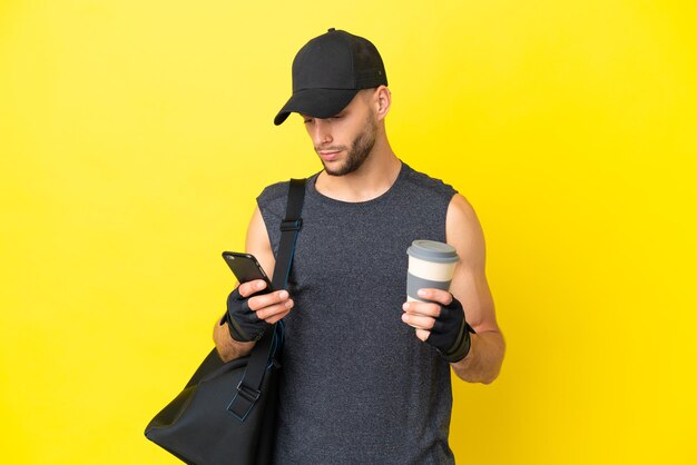 Młody sportowy blondyn ze sportową torbą na białym tle na żółtym tle, trzymający kawę na wynos i telefon komórkowy
