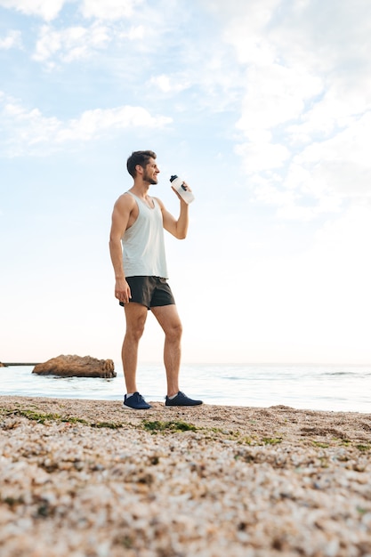 Młody sportowy sportowiec odpoczywa po joggingu na plaży i pije wodę