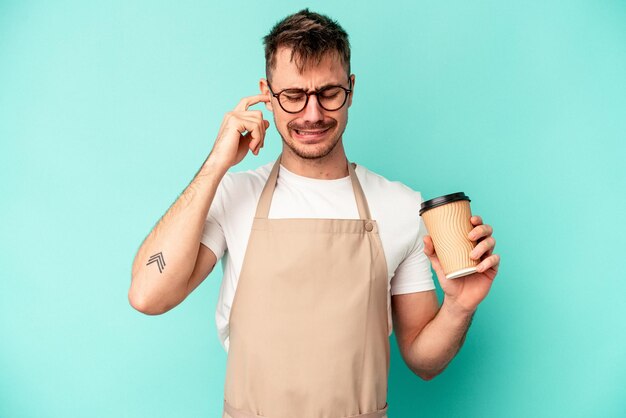 Młody sprzedawca mężczyzna trzyma kawę na białym tle na niebieskim tle obejmujące uszy rękami.