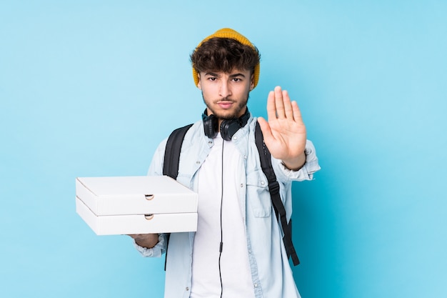 Młody student arabski mężczyzna trzyma pizze na białym tle stojący z wyciągniętą ręką pokazuje znak stopu, uniemożliwiając ci.