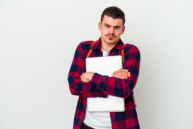 Młody student kaukaski mężczyzna trzyma laptop na białym tle na białej ścianie marszcząc brwi z niezadowoleniem, trzyma założone ręce.