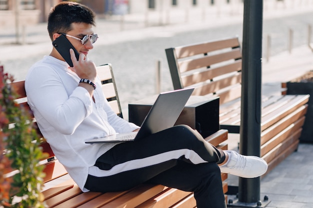 Młody stylowy facet w koszuli z telefonu i notebooka na ławce w słoneczny ciepły dzień na zewnątrz, niezależny