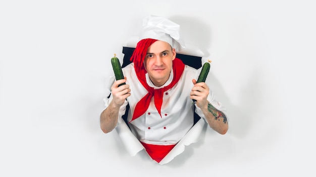 Młody szalony mężczyzna ubrany jak szef kuchni z ogórkami Szalony kucharz z warzywami wystającymi z dziury na białym tle