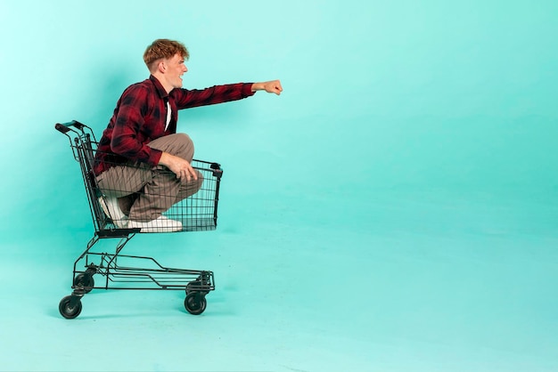 Młody szczęśliwy kupiec siedzi i jeździ w wózku z supermarketu na niebieskim tle