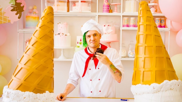 Młody szczęśliwy mężczyzna przebrany za szefa kuchni z dredami jedzący babeczkę w jasnej cukierni