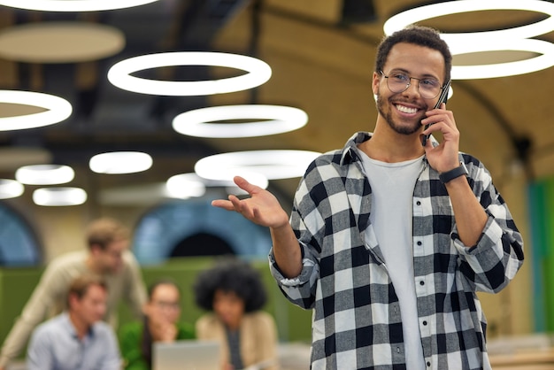 Młody szczęśliwy mężczyzna rasy mieszanej rozmawia przez telefon i uśmiecha się stojąc w przestrzeni coworkingowej