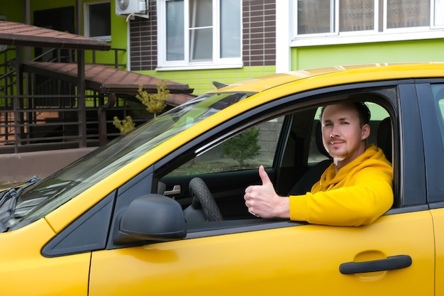 Młody szczęśliwy mężczyzna taksówkarz siedzi za kierownicą taksówki i pokazuje jak
