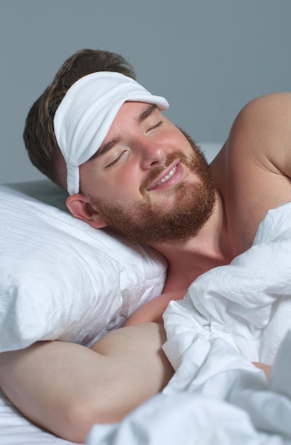 Młody szczęśliwy przystojny śpiący mężczyzna w łóżku w sypialni w domu rano, leżąc pod białym kocem, dobrze odpoczywa w nocnej masce na twarzy zdjęcie pionowe