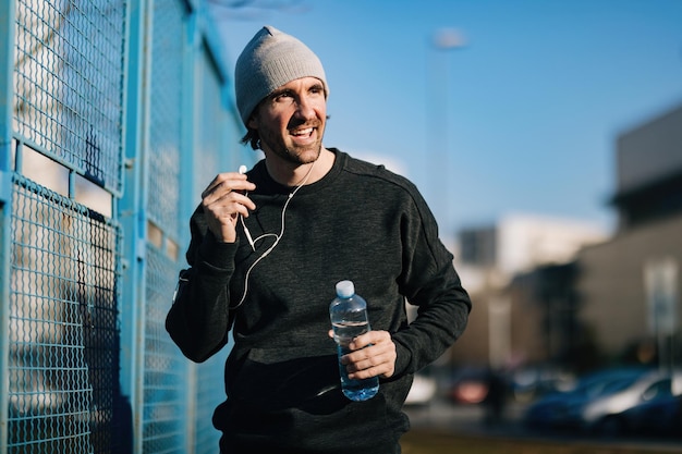 Młody szczęśliwy sportowiec niosący butelkę wody i dopasowujący słuchawki podczas ćwiczeń w mieście