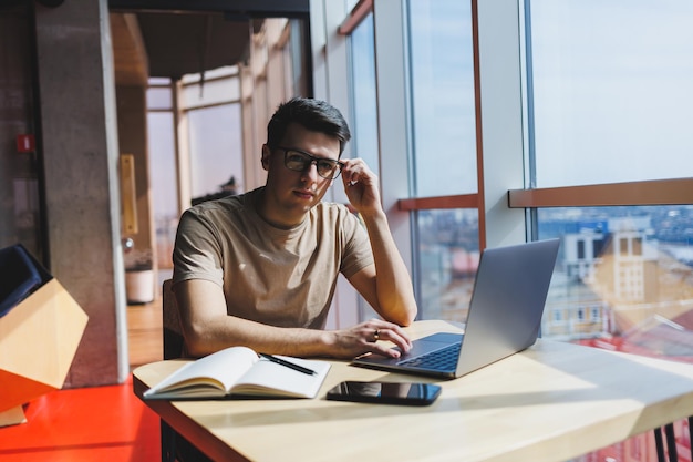 Młody tysiącletni mężczyzna siedzący przy stole z przodu otwartego laptopa podczas nauki online Nowoczesna cyfrowa praca nomadów na freelance na netbooku we wnętrzu kawiarni