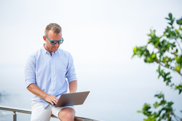 Zdjęcie młody udany biznesmen mężczyzna pracuje z laptopem na wakacjach nad morzem. nosi okulary przeciwsłoneczne, koszulę i białe szorty. praca poza biurem, freelancer