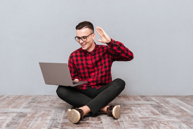 Młody uśmiechający się hipster w okularach, siedząc na drewnianej podłodze z laptopa i macha w aparacie laptopa. Na białym tle szarym tle