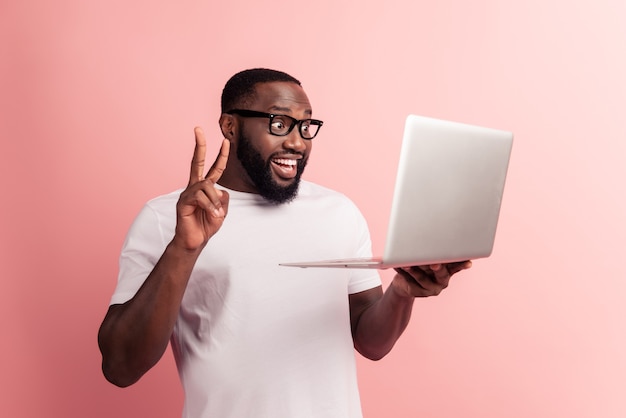 Młody uśmiechnięty afrykański mężczyzna stojący i korzystający z laptopa sprawia, że zdalne wideorozmowa pokazuje znak v