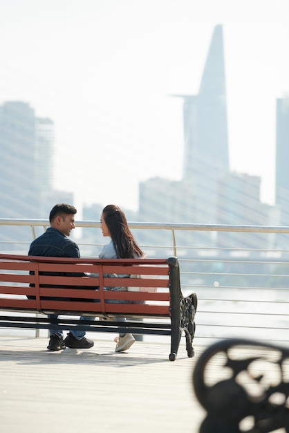 Młody uśmiechnięty chłopak i dziewczyna siedzą na ławce naprzeciwko miasta i rozmawiają o swoim związku