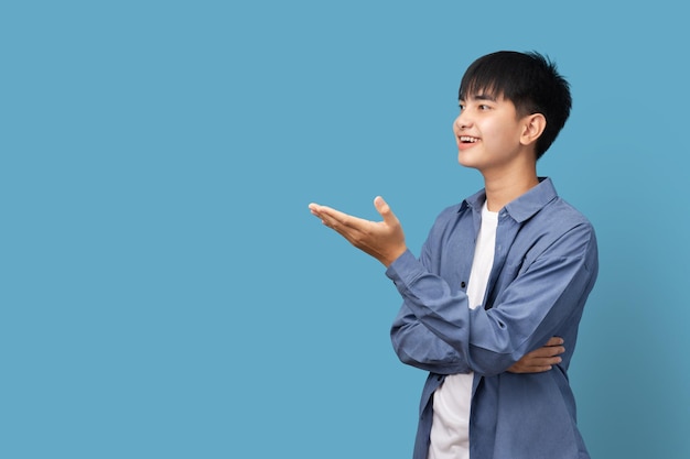 Młody uśmiechnięty mężczyzna pokazuje wyciągniętą rękę z palmą, azjatycki mężczyzna stojący na niebieskim tle, miejsce na produkt lub tekst.