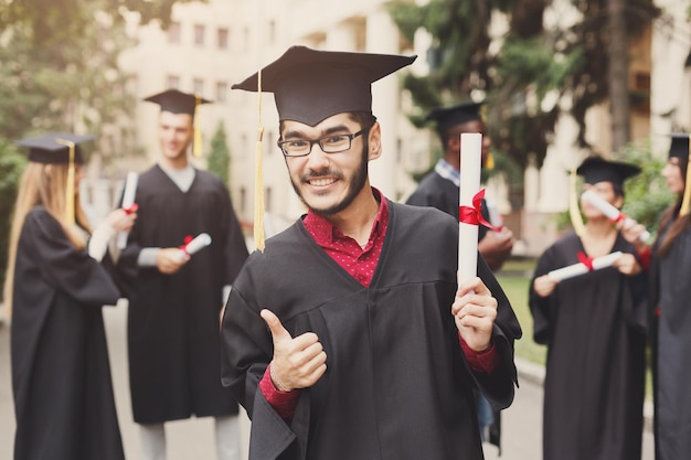 Młody Uśmiechnięty Mężczyzna W Dniu Ukończenia Studiów Pokazując Kciuk Do Góry Stojący Z Wieloetniczną Grupą Studentów. Koncepcja Edukacji, Kwalifikacji I Sukni.