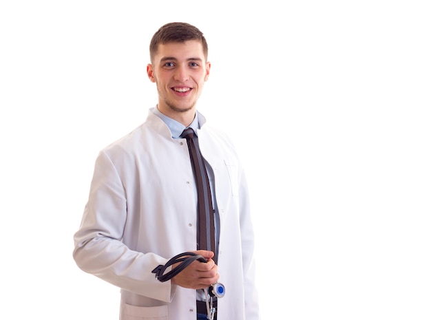 Młody uśmiechnięty mężczyzna z brązowymi włosami w niebieskiej koszuli krawat i suknia lekarska, trzymający stetoskop w studio