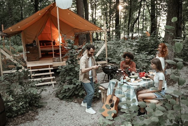 Zdjęcie młody uśmiechnięty mężczyzna zabawiający przyjaciół grających w kendamę na glamping, ciesząc się imprezą przy grillu. mecz pucharu i piłki. drewniana zabawka. wielorasowi milenialsi spędzają czas na biwaku z gitarą i piknikiem