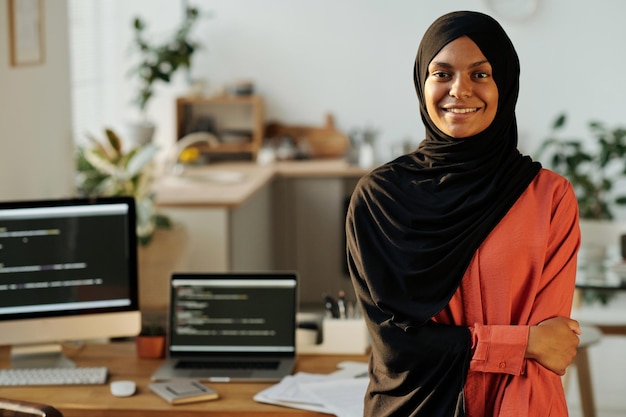 Młody uśmiechnięty muzułmański pracownik domowego biura w czarnym hidżabie i czerwonej koszuli patrzący na kamerę