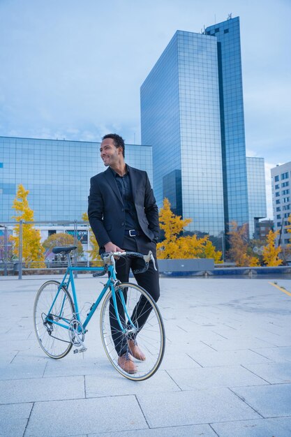 Młody, uśmiechnięty, stylowy biznesmen jedzie do biura na rowerze przed dzielnicą finansową.