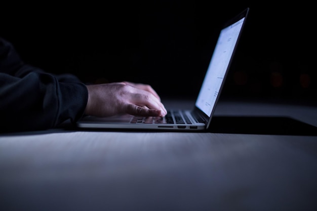 Młody utalentowany haker korzystający z laptopa podczas pracy w ciemnym biurze z dużymi światłami miasta w tle w nocy