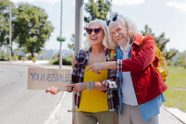 Młody w duszy. Pozytywny starszy mężczyzna, wskazując na znak podczas autostopu razem z żoną