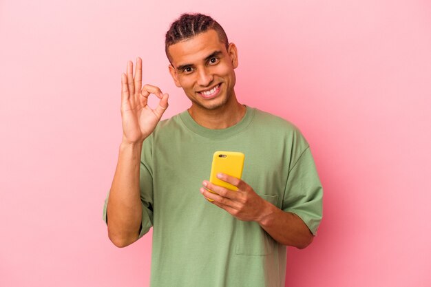 Młody wenezuelski mężczyzna trzymający telefon komórkowy na białym tle na różowym tle wesoły i pewny siebie pokazując ok gest.