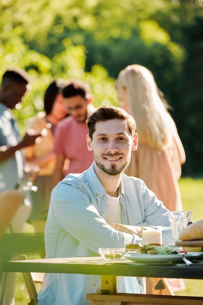 Młody Wesoły Kaukaski Mężczyzna W Casual, Siedzący Przy Stole Na świeżym Powietrzu, Podczas Gdy Jego Przyjaciele Razem Gotują Grill