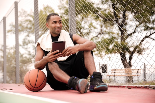 Młody wesoły koszykarz siedzi w parku w pobliżu koszykówki i trzymając tablet