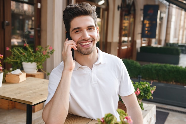 młody wesoły mężczyzna siedzi w kawiarni na świeżym powietrzu, rozmawiając przez telefon komórkowy