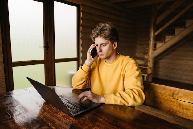 Młody wolny strzelec pracuje w drewnianym wiejskim domu na laptopie przy stole
