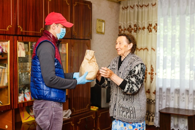 Młody wolontariusz w masce daje starszej kobiecie pudełka z jedzeniem w pobliżu jej domu. Syn pomaga samotnej starszej matce. Wsparcie rodziny, opieka. Poddany kwarantannie, odizolowany. Koronawirus (COVID-19. Darowizna