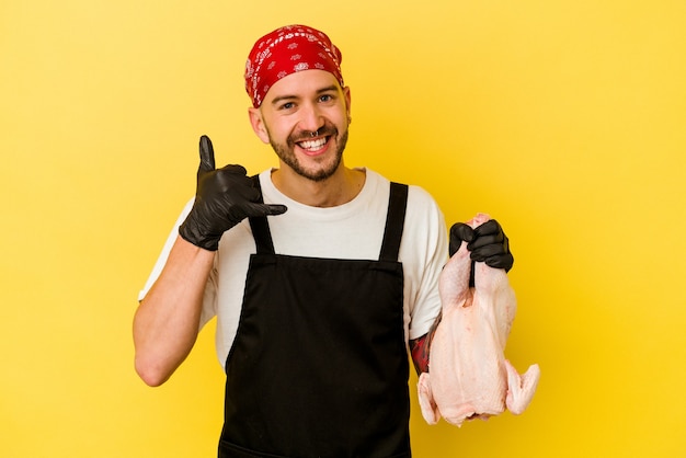 Młody wytatuowany dozownik kaukaski mężczyzna trzyma kurczaka na białym tle na żółtym tle przedstawiający gest telefonu komórkowego palcami.