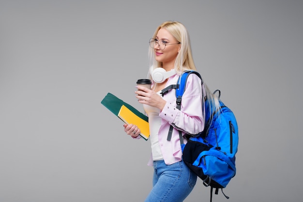 Młody żeński uczeń w hełmofonach z plecakiem pije kawę, ono uśmiecha się. Uruchomienie, freelancer. Nauka języków obcych online, przygotowanie do egzaminu