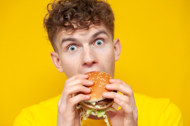 Młody zszokowany facet zjada dużego burgera i jest zaskoczony na żółtym tle mężczyzna gryzie fast food zbliżenie