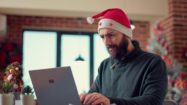 Młodych dorosłych pomysłów na burzę mózgów, aby stworzyć raport na laptopie, nosząc czapkę Świętego Mikołaja w biurze z choinką. Świąteczny mężczyzna pracujący na komputerze z danymi badawczymi w okresie wakacyjnym. Strzał z ręki.