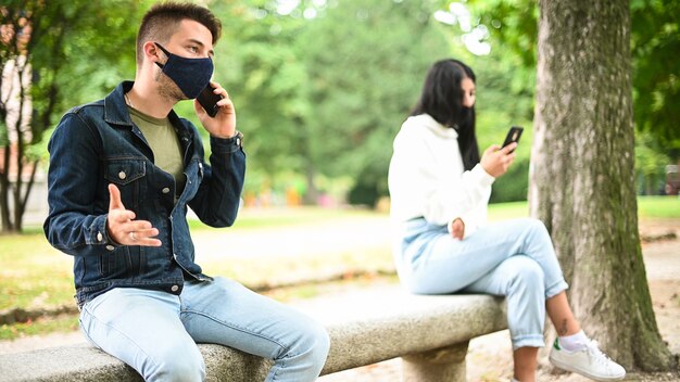 Młodzi ludzie zachowujący dystans społeczny z powodu koronawirusa podczas korzystania z telefonu na świeżym powietrzu w parku