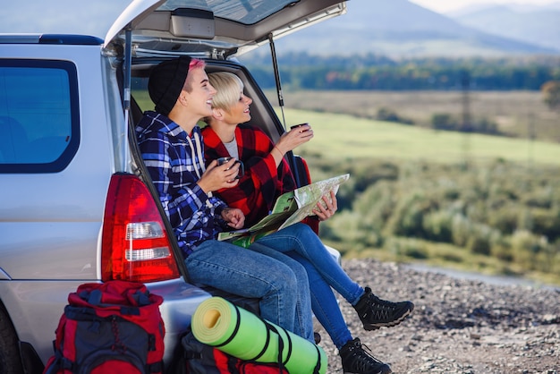 Młodzi przyjaciele podróżujący siedzą na bagażniku samochodu z czapką herbaty i patrzą na papierową mapę.