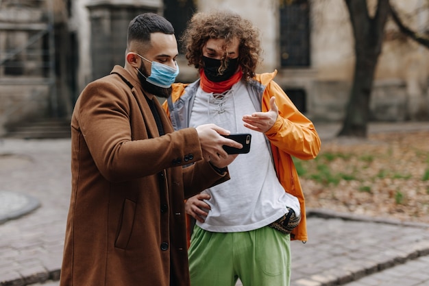 Młodzi przystojni mężczyźni w maskach medycznych za pomocą nowoczesnego smartfona, stojąc na ulicy. Szczęśliwi faceci spacerujący na świeżym powietrzu w czasie wolnym.