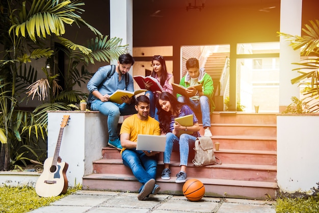 Młodzi studenci z Azji i Indii czytający książki, uczący się na laptopie, przygotowujący się do egzaminu lub pracujący nad projektem grupowym siedząc na trawie, schodach lub stopniach kampusu uniwersyteckiego