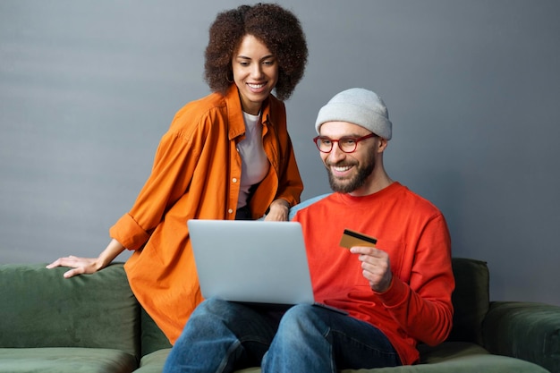 Młodzi uśmiechnięci przyjaciele używający laptopa trzymającego kartę kredytową na zakupy online siedząc w domu