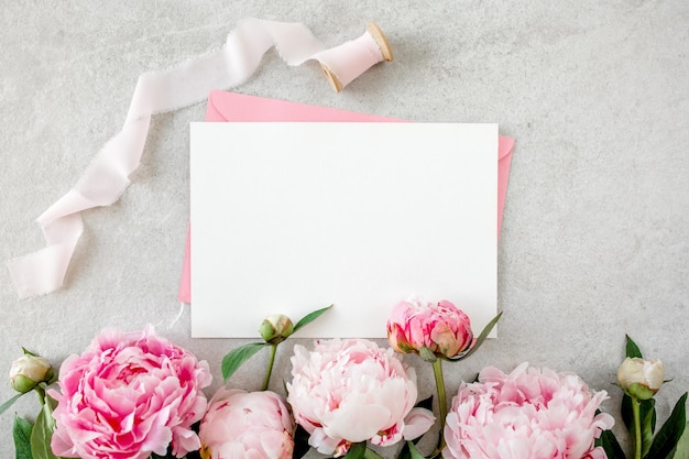 Zdjęcie mockup zaproszenia pusta karta powitalna różowa koperta i piwony na szarym kamiennym stole kwiatowe tło płaski widok z góry