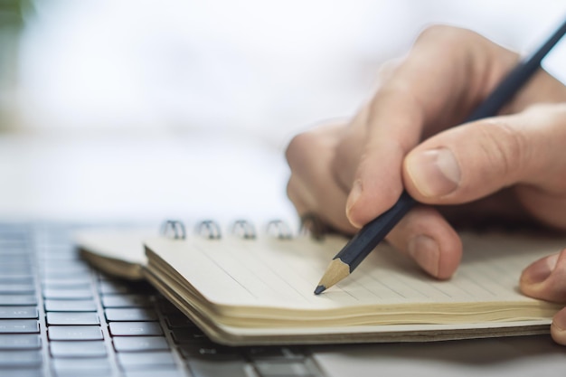 Zdjęcie mocno skup się na dłoni mężczyzny, gdy pisze w notatniku na subtelnie rozmytym tle współczesnego laptopa