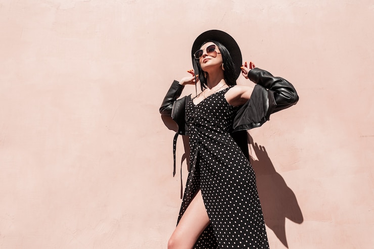Zdjęcie moda czarowny młoda kobieta w stylowe okulary w skórzanej kurtce w pięknej sukni prostuje kapelusz w słoneczny dzień. urocza modna dziewczyna w eleganckich czarnych letnich ubraniach cieszy się promieniami słonecznymi na zewnątrz.