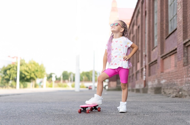 Zdjęcie moda dziewczynka w okularach przeciwsłonecznych z różową deskorolką w mieście