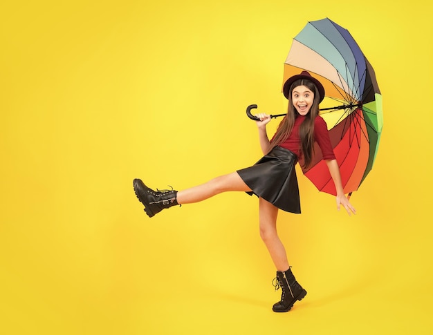 Moda jesienna pogoda dla dzieci Dziecko nastolatek dziewczyna z parasolem jesienią czarna skórzana spódnica buty i kapelusz na żółtym tle studyjny Podekscytowana nastolatka zadowolona zdumiona i uradowana emocjami