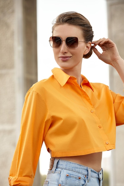 Moda na zewnątrz portret eleganckiej luksusowej kobiety w czarnych okularach przeciwsłonecznych modna pomarańczowa koszula i dżinsy Skopiuj puste miejsce na tekst