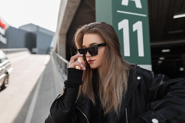 Moda piękna kobieta w modnych okularach przeciwsłonecznych w stylowej skórzanej kurtce z czarną bluzą z kapturem spaceruje po mieście w pobliżu parkingu