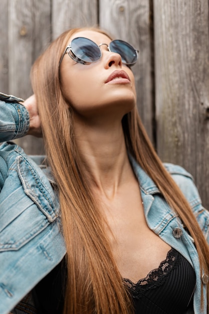Moda portret glamour ładnej modelki w niebieskich stylowych okrągłych okularach przeciwsłonecznych z dżinsową kurtką i czarną koronkową bielizną w pobliżu drewnianej ściany