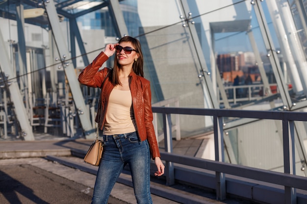 Moda portret młodej modelki w brązowej skórzanej kurtce, dżinsach i okularach przeciwsłonecznych na wielkomiejskim.